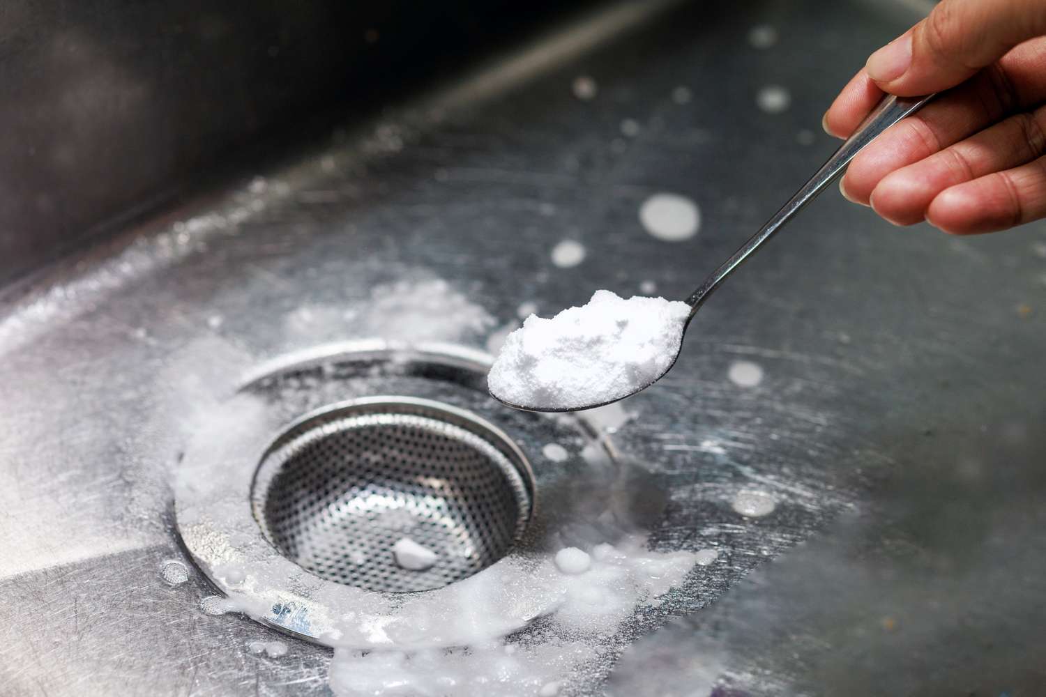 hand holding spoon with baking powder over a stainless steel sink and drian