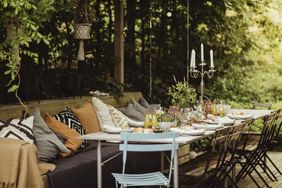 An outdoor dining table set with food drinks and candles under a shaded leafy area