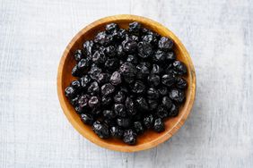 A wooden bowl filled with dried black berries on a light textured background