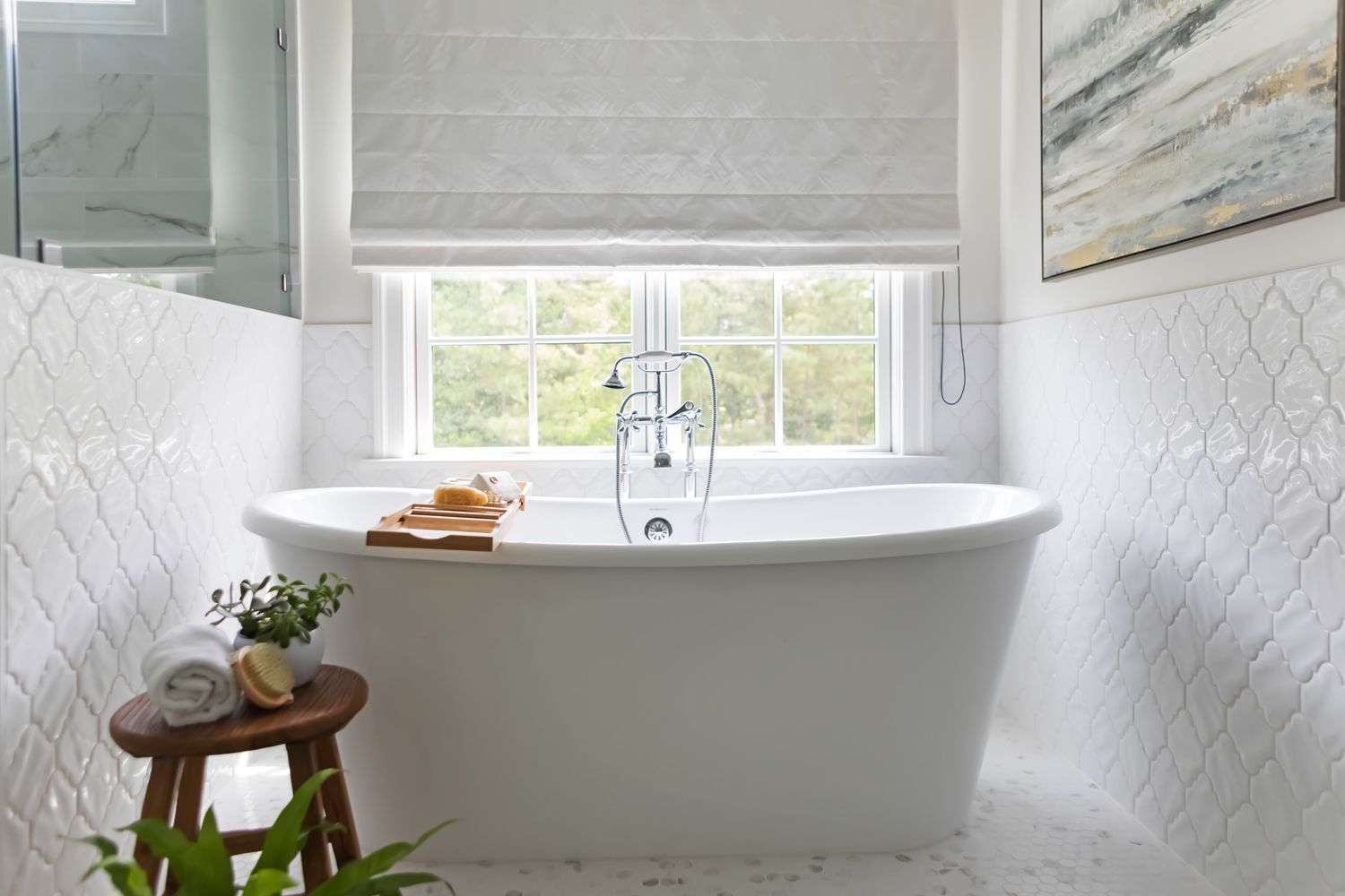 Freestanding bathtub in a bathroom with a small side table holding plants and towels, a window with roman shade above