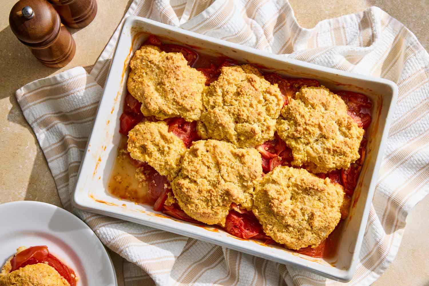Baking dish filled with tomato cobbler layered tomatoes and biscuit topping placed on a striped kitchen towel