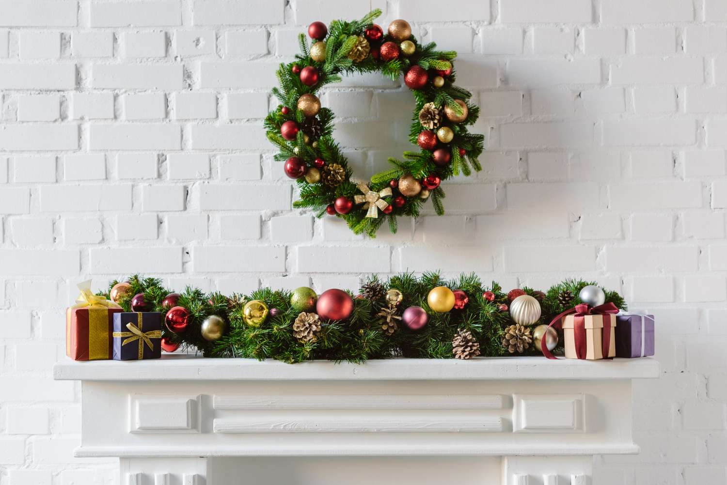A fireplace mantel decorated with garlands ornaments and wrapped presents with a festive wreath hanging above set against a white brick wall