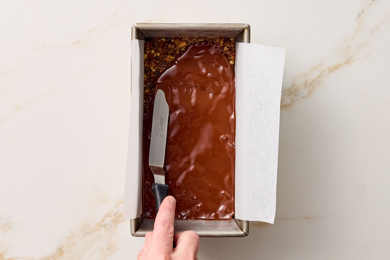 A persons hand using a knife to spread melted chocolate on a date nut bar in a baking pan