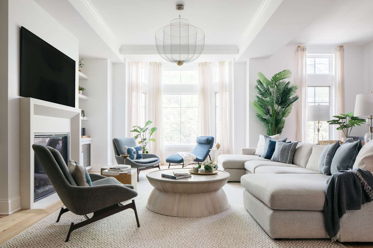 Minimalist living room by Outline Interiors: beige sectional sofa, blue accent chairs, wood coffee table with books, stone fireplace, lush potted plants, TV, and large windows with sheer curtains. Light wood floor and natural light create a calm, stylish space.