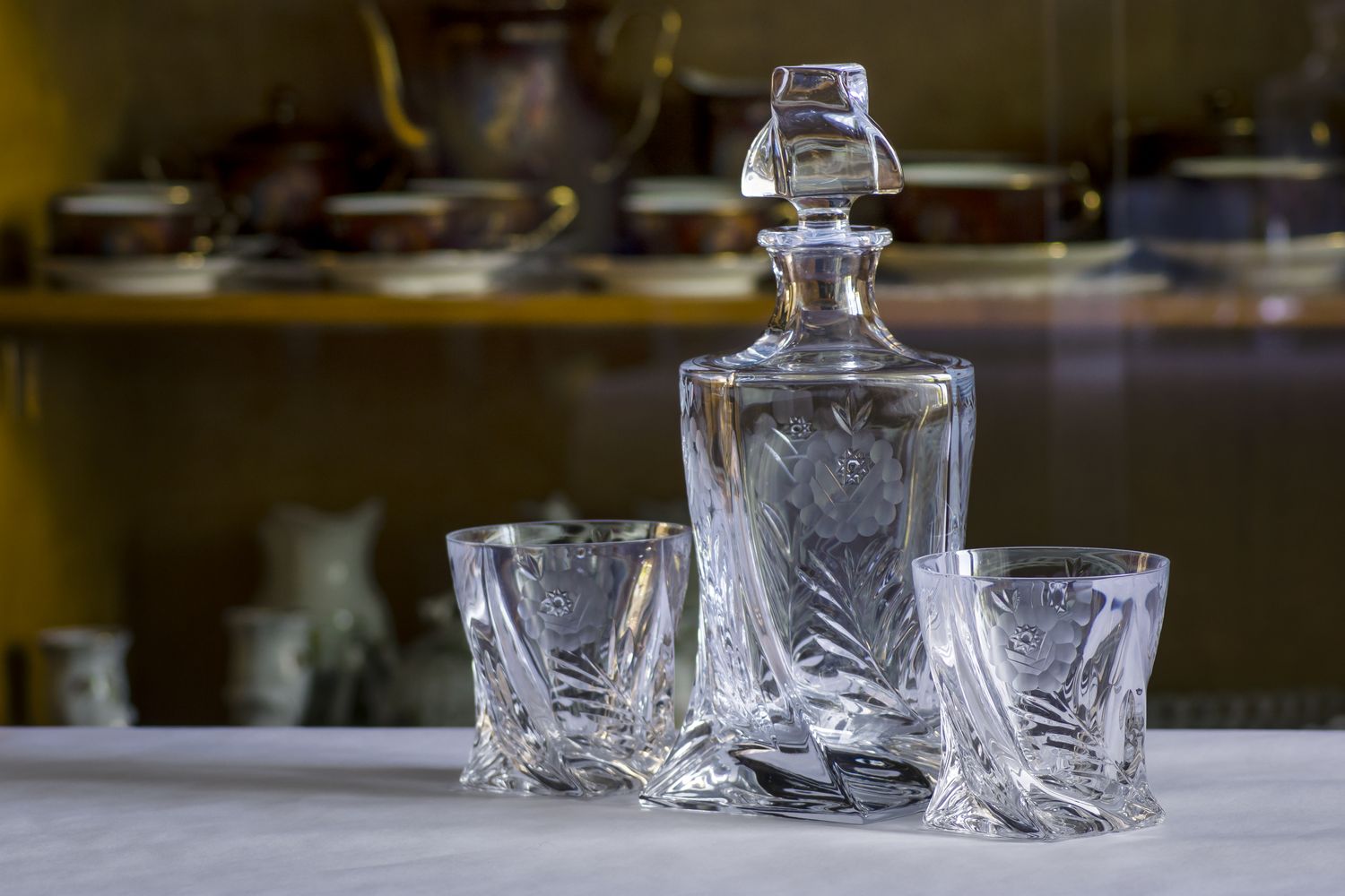 Crystal decanter with two matching glasses on a table background showing a cabinet with dishes