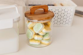 A glass jar with wooden lid containing stacked cleaning tablets on a surface with other storage containers