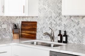 A modern kitchen featuring a double sink herringbone tile backsplash a knife block bottles on the counter and a wooden cutting board