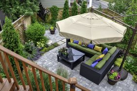 A backyard patio with seating area under a large umbrella surrounded by plants and a wooden fence