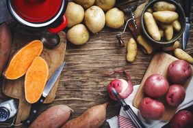 raw sweet potatoes and 3 types of white potatoes on wood surface with peeler and knives