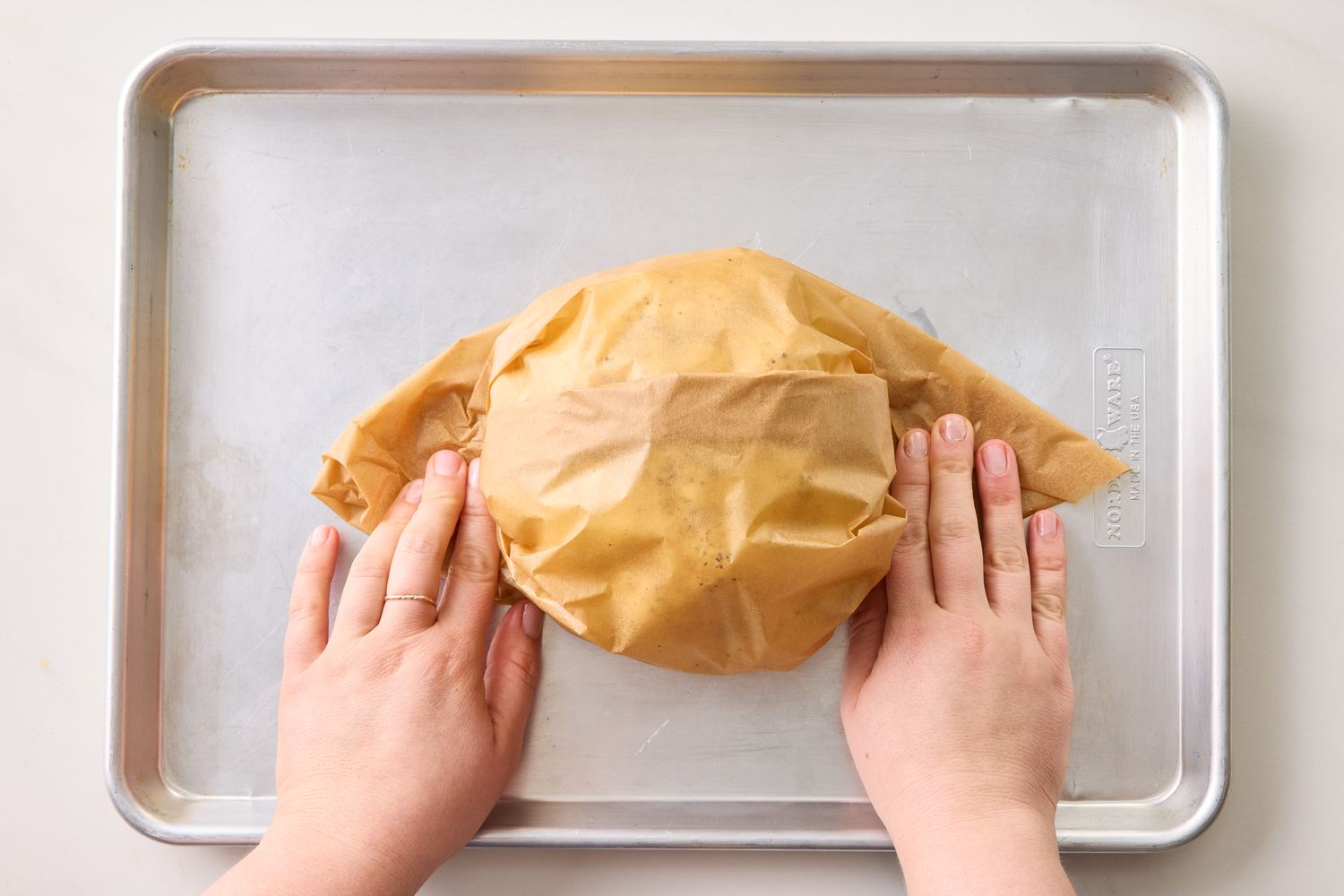Hands wrapping a head of cauliflower in parchment paper on a baking sheet