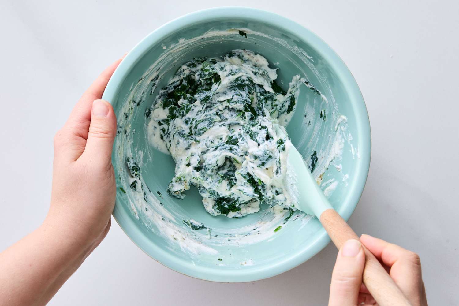 Hands mixing spinach dip in a bowl with a spatula