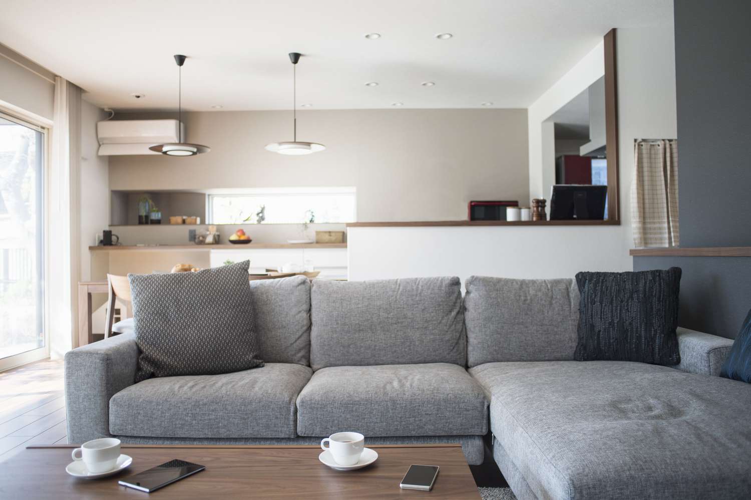 A modern living room with a gray couch a wooden coffee table with cups and a phone and an open dining area in the background