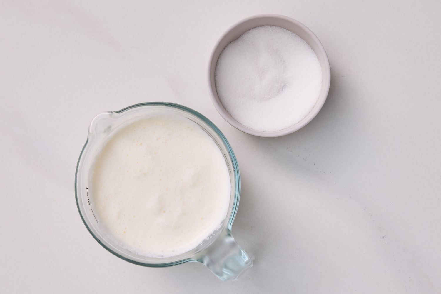 A measuring cup of heavy cream next to a small bowl containing sugar suitable for making whipped cream