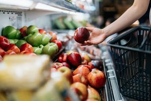 A shopper selecting an apple from a display of various fruits holding a basket at a grocery store