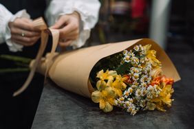 A floral bouquet being tied with a ribbon by a person dressed in formal attire featuring daisies and other yellow flowers
