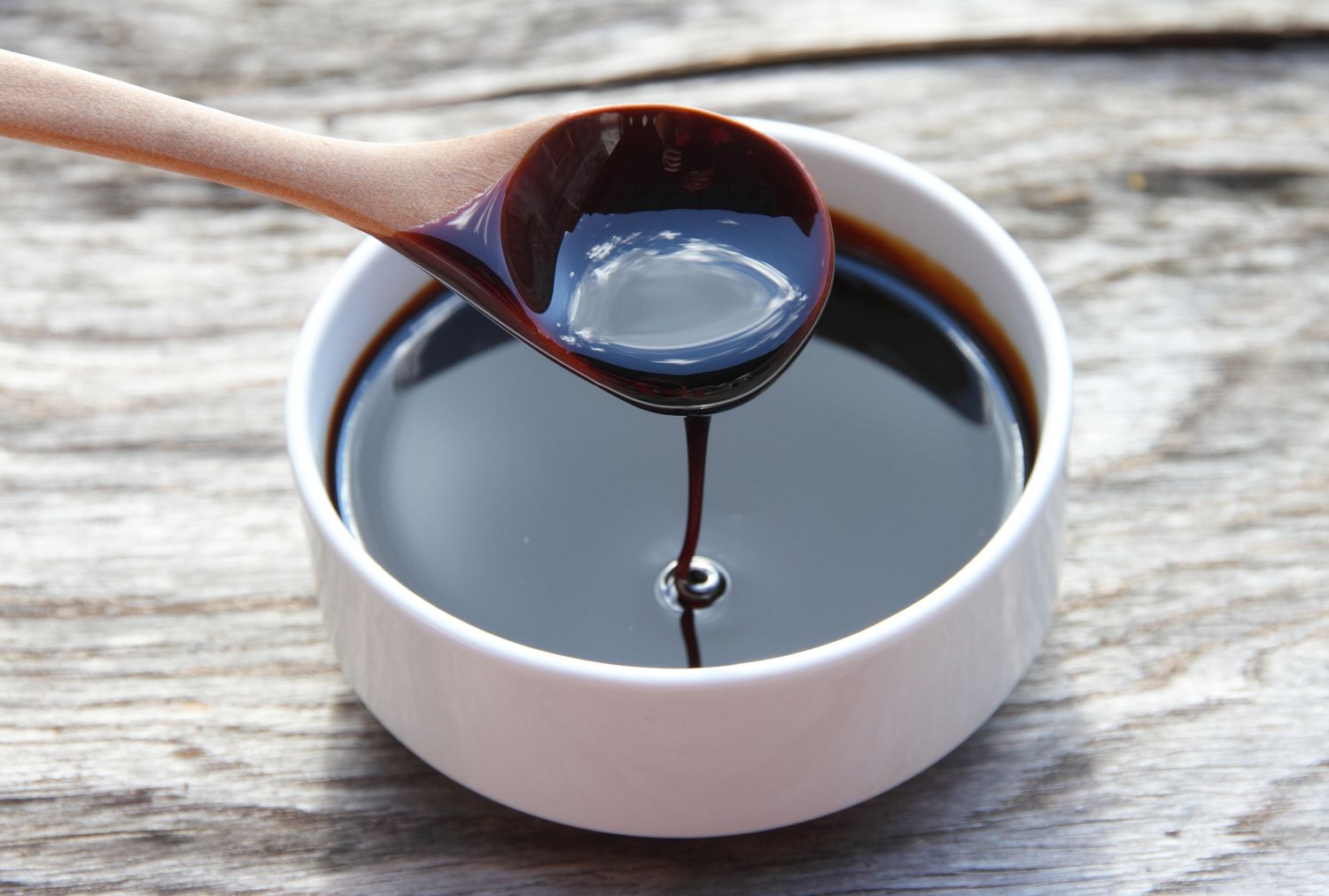 A wooden spoon pouring molasses into a white bowl on a wooden surface