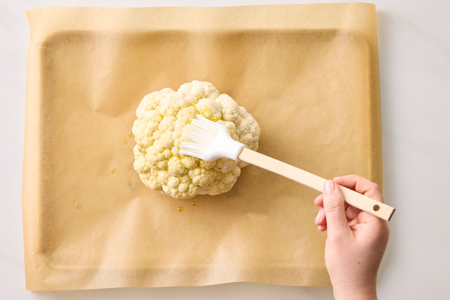 A hand brushing oil onto a whole cauliflower placed on parchment paper