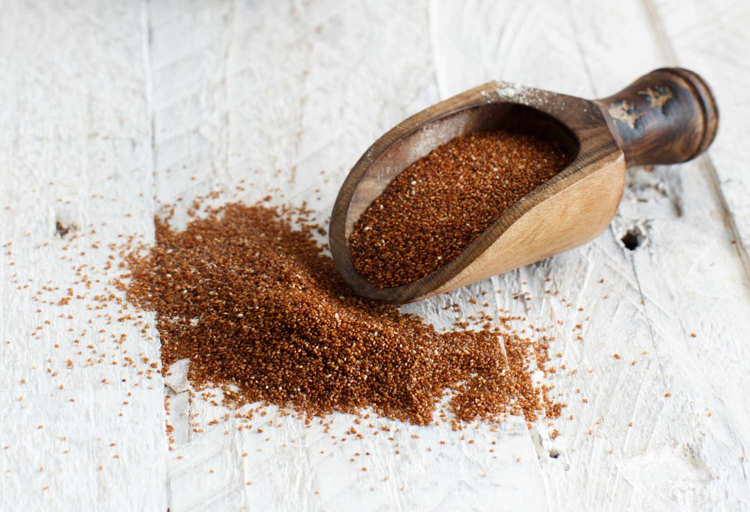 uncooked teff in a wooden scoop and on a white surface