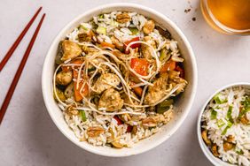 A bowl of rice topped with vegetables noodles and pieces of chicken accompanied by chopsticks and a drink
