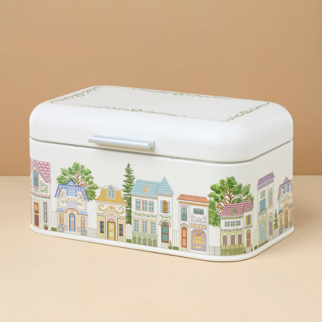 A rectangular tin container with painted images of colorful houses and trees on its sides resting on a flat surface