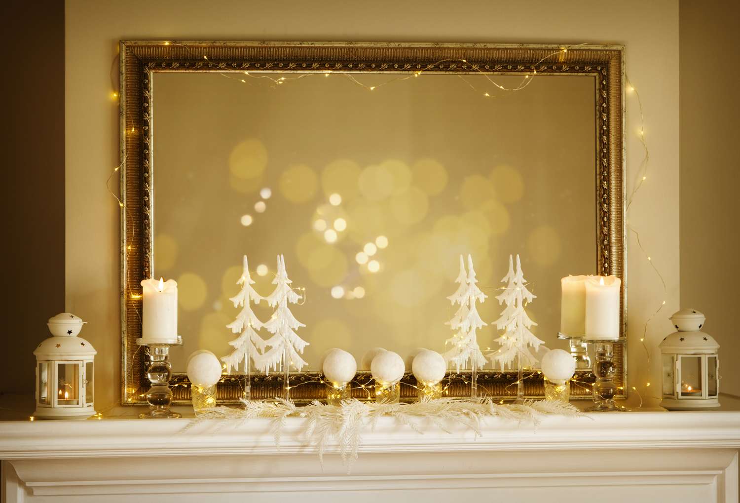 A mantel decorated with candles ornaments and miniature trees with a lighted mirror in the background