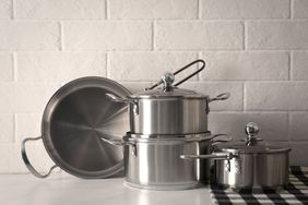 A set of stainless steel cookware including pots and pans arranged on a countertop with a brick wall in the background