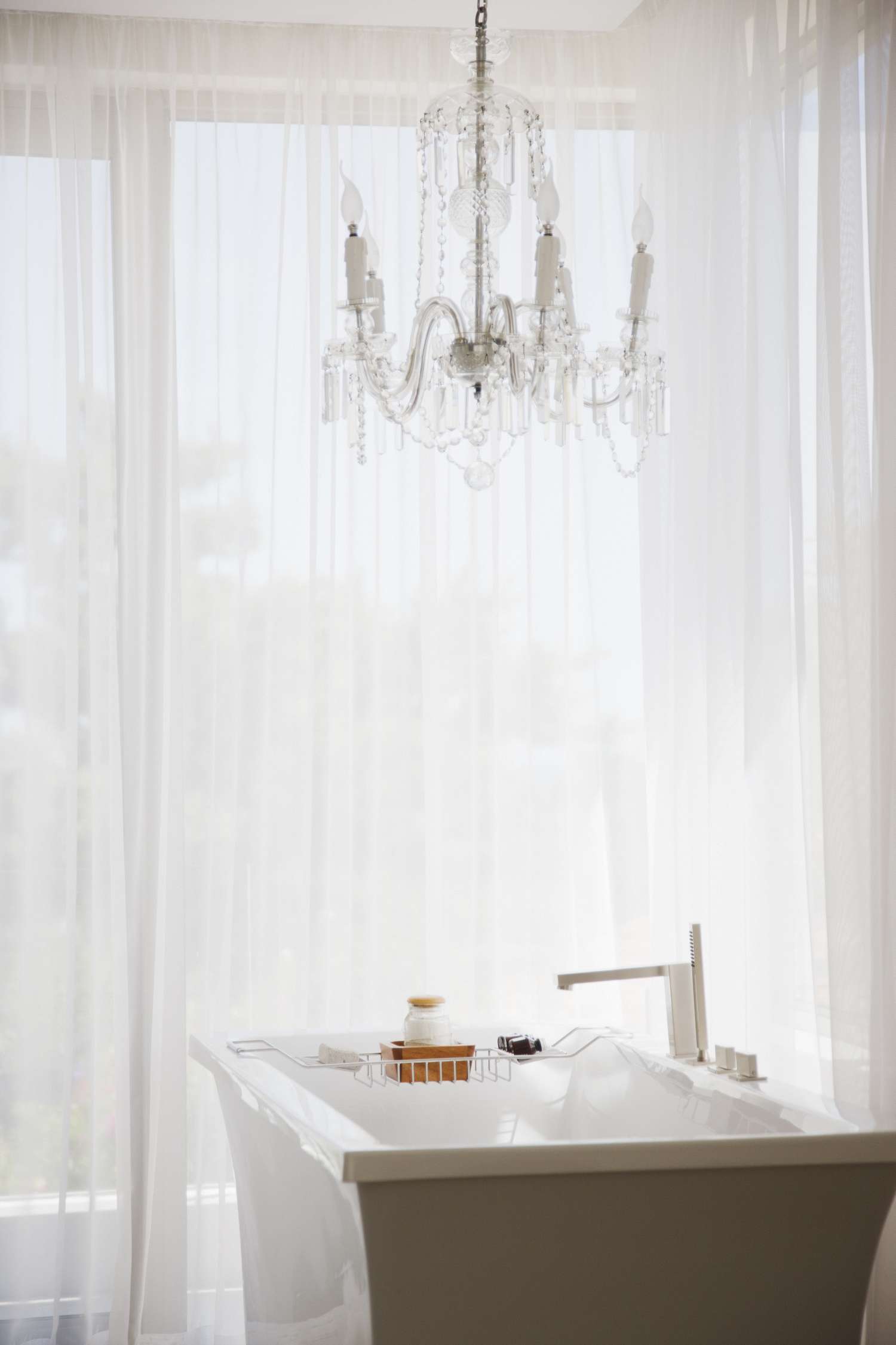 A freestanding bathtub in a room with sheer curtains featuring a chandelier above