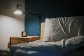 A bedroom scene showing a lit wall lamp above a nightstand with a small clock adjacent to a bed with pillows and a blanket