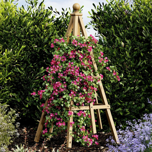 A wooden trellis supporting blooming pink flowers in a garden surrounded by bushes and other plants