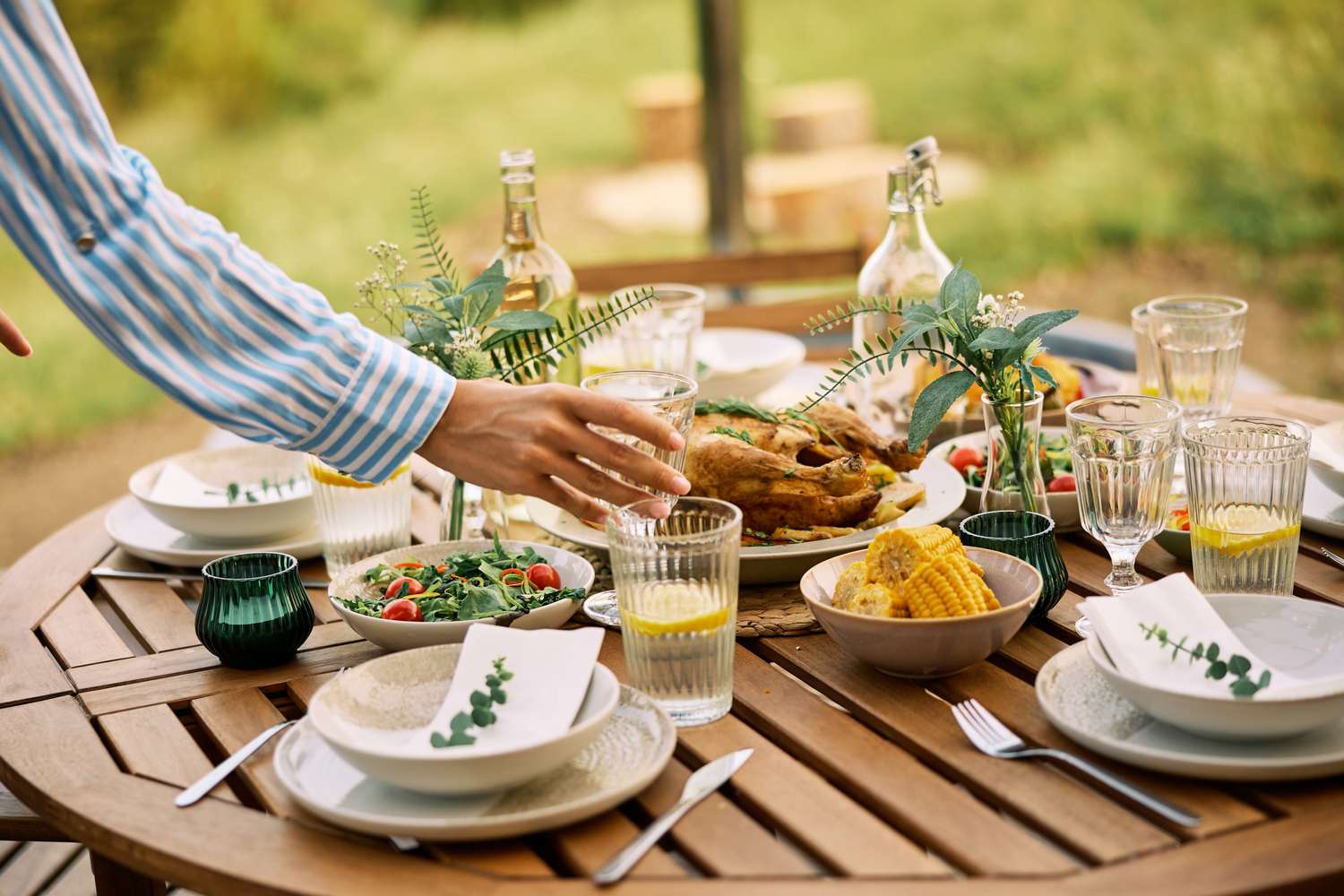 A wooden outdoor table set with dishes glasses floral decorations and food including corn and salad with a person reaching to arrange items