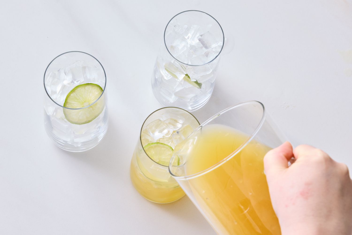 A hand pours a pineapple rum cocktail into a glass with ice surrounded by glasses with lime slices and ice on a table