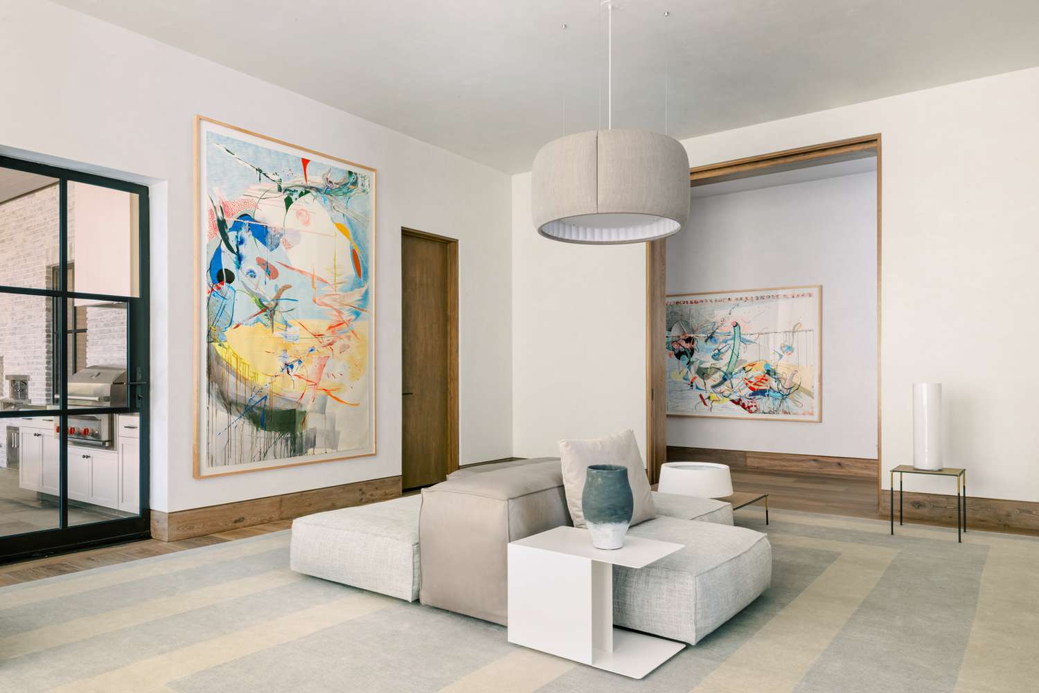 Minimalist living room by Lambrakos Studio: modular gray upholstered seating with beige cushions, white side table, vibrant abstract art on walls, fabric pendant light, and striped rug. Large windows, wood floor, and natural light create a calm, stylish space.