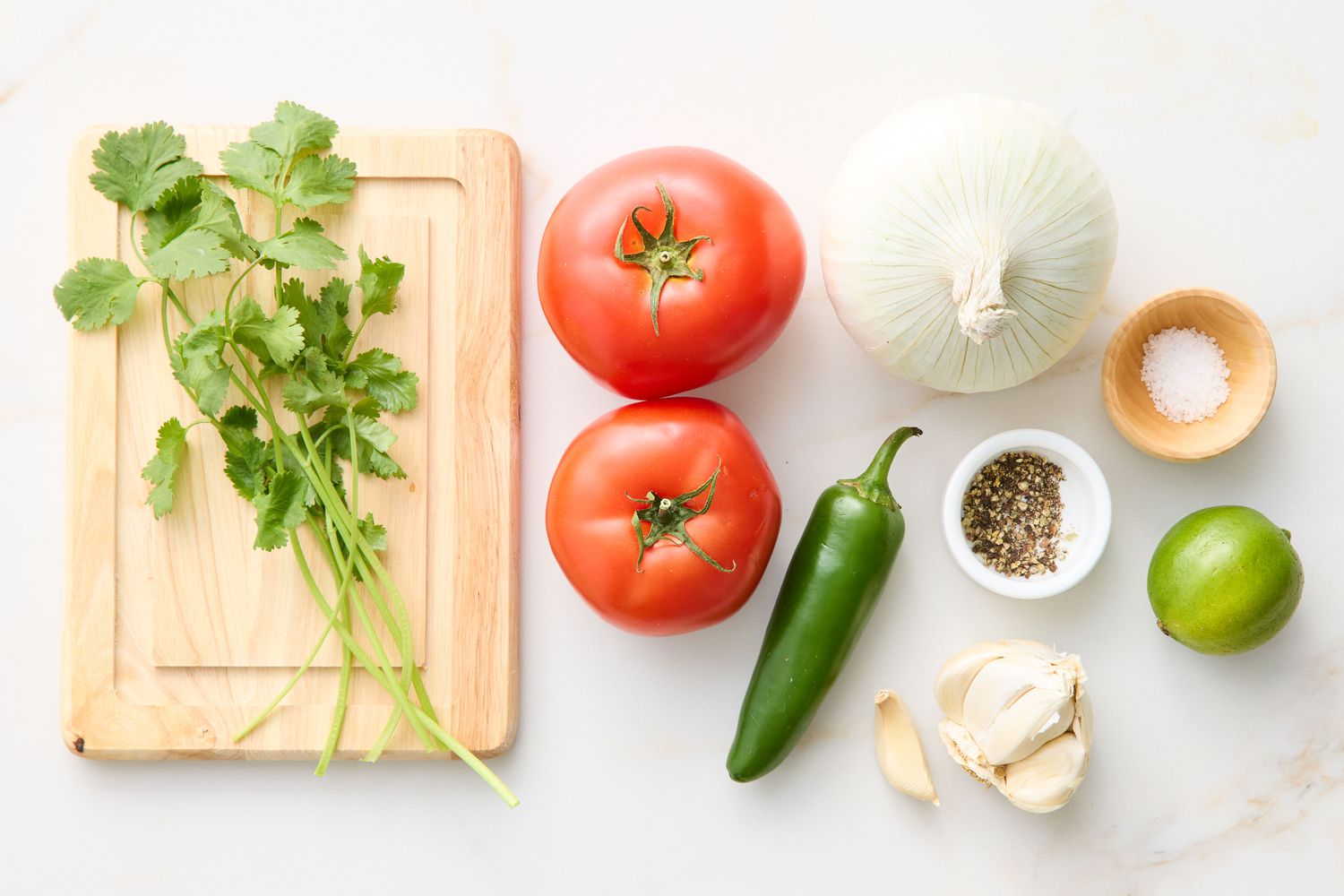 Ingredients for a salsa recipe including cilantro, tomatoes, onion, jalapeño, garlic, lime, salt, and spices arranged on a surface