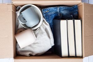 A cardboard box containing stacked books folded jeans a cloth and two ceramic mugs