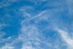 A crescent moon visible in a daytime sky scattered with clouds