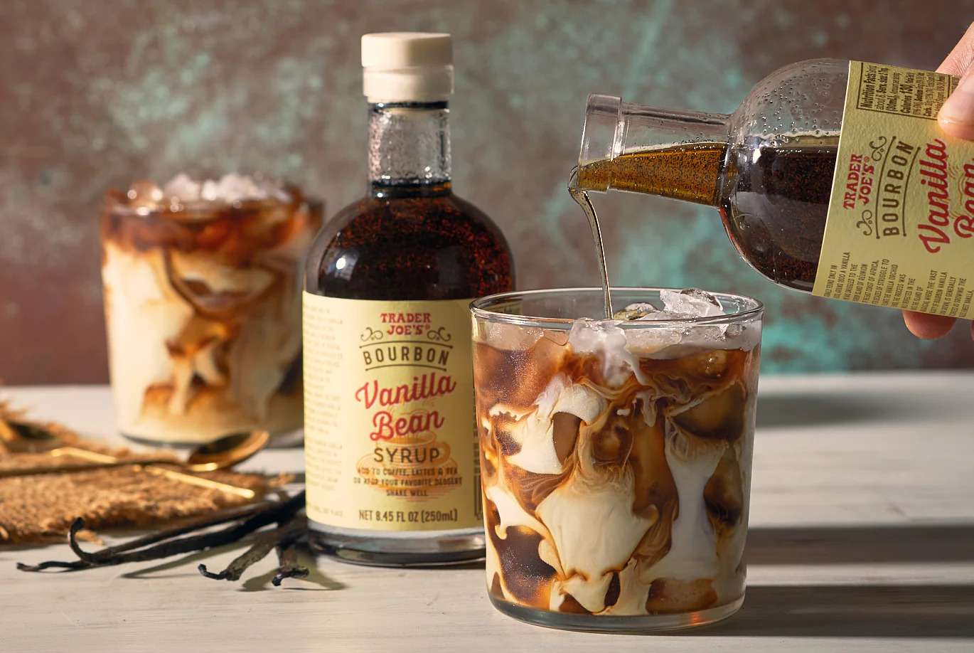 An iced beverage being prepared with vanilla bean syrup with syrup bottles and glasses on a table