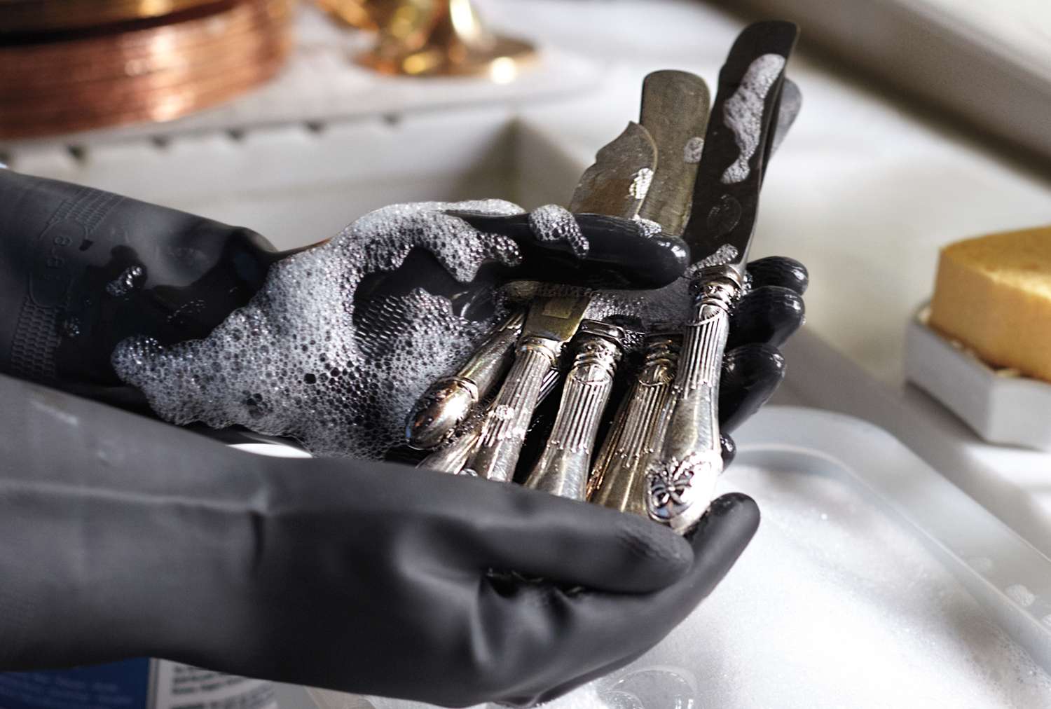 Hands wearing gloves cleaning silver utensils with soap suds