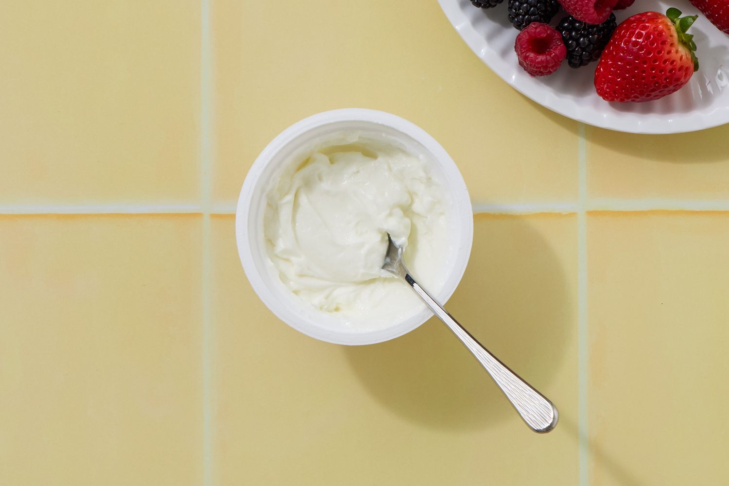 A container of Greek yogurt with a spoon placed next to a plate of strawberries and other berries on a tiled surface
