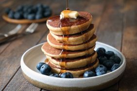 stack of fluffy pancakes on white plate with butter, syrup, and blueberries