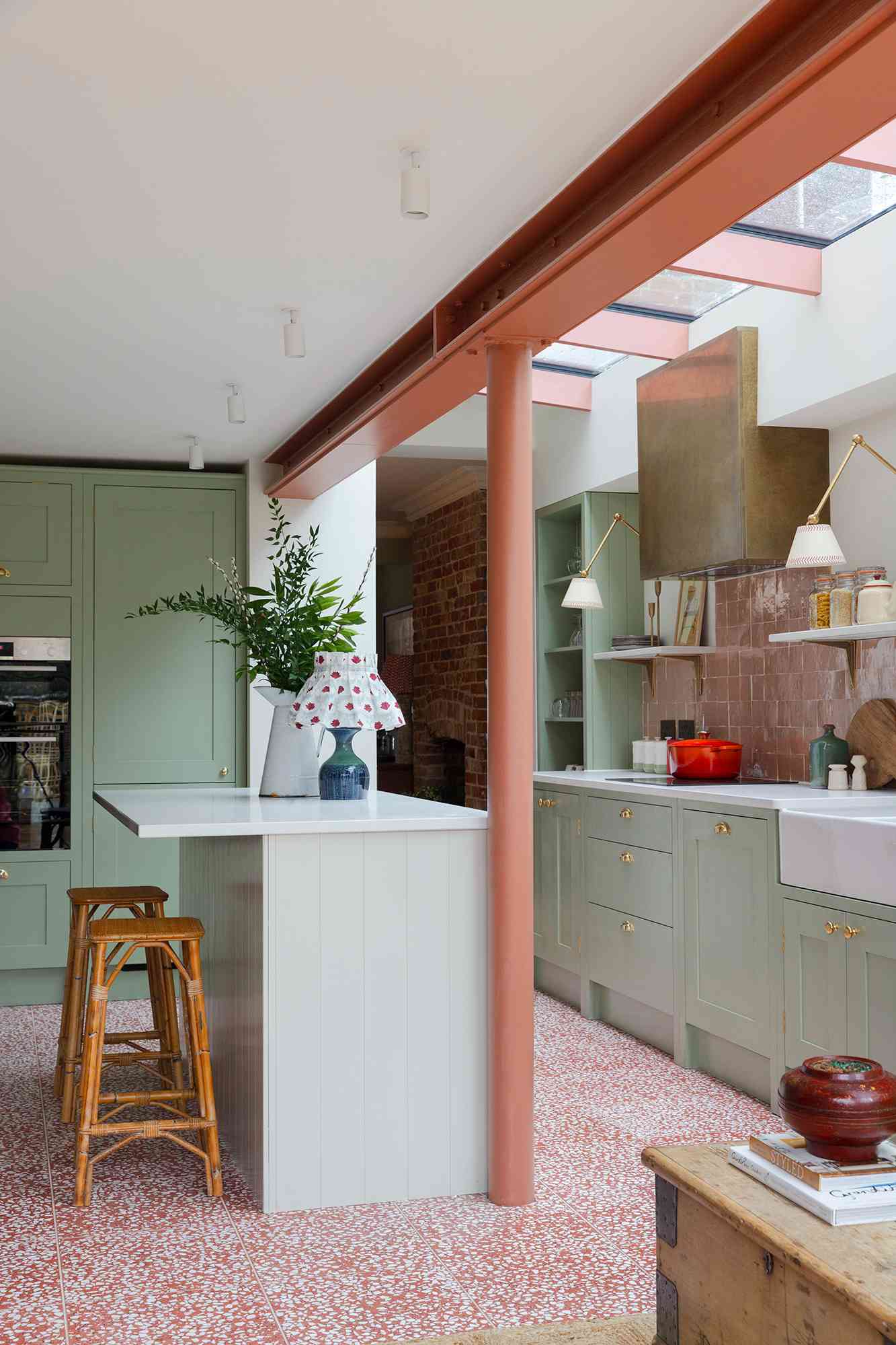 Kitchen design by Otto Tiles & Design and Vaughan Design & Development | Photography by Chris Snook: soft sage cabinetry, brass hardware, warm terracotta accents, and pink terrazzo flooring. A bold brass range hood and vintage-inspired lighting complete the eclectic, modern look.