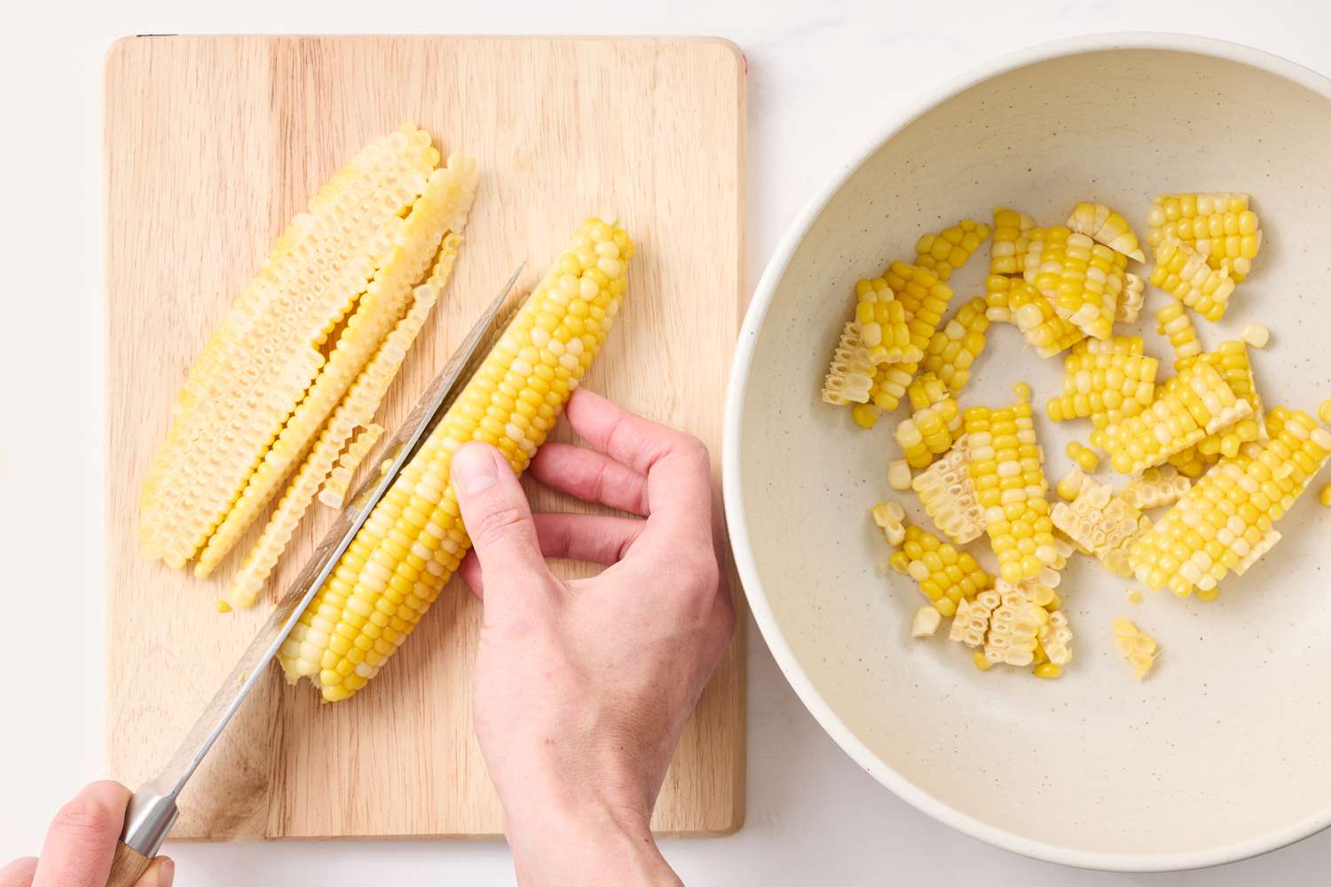 A person cutting corn kernels off a cob onto a cutting board with a bowl containing the cut kernels nearby