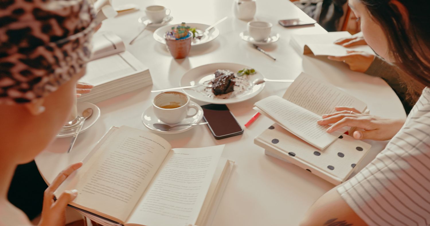 People sitting around a table reading books cups and plates are on the table