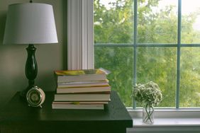 Stack of books on a table with a lamp and flowers near a window