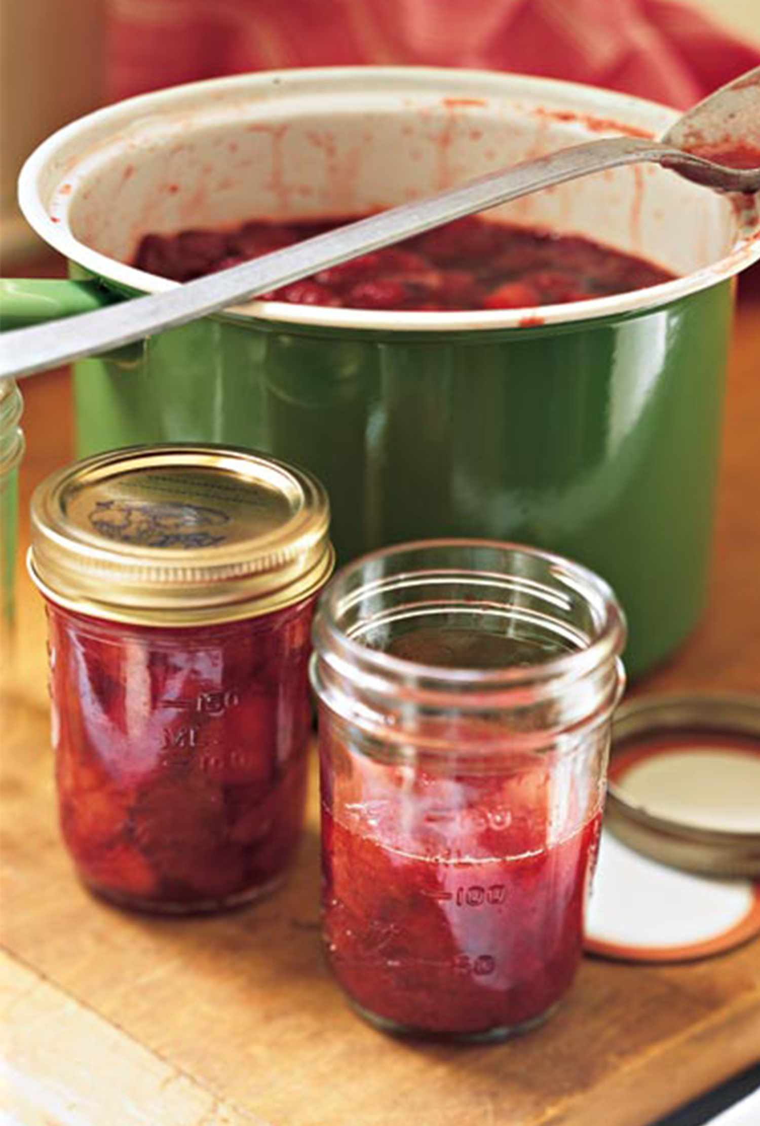 Jars of strawberry preserves next to a green pot filled with preserves with a ladle resting inside