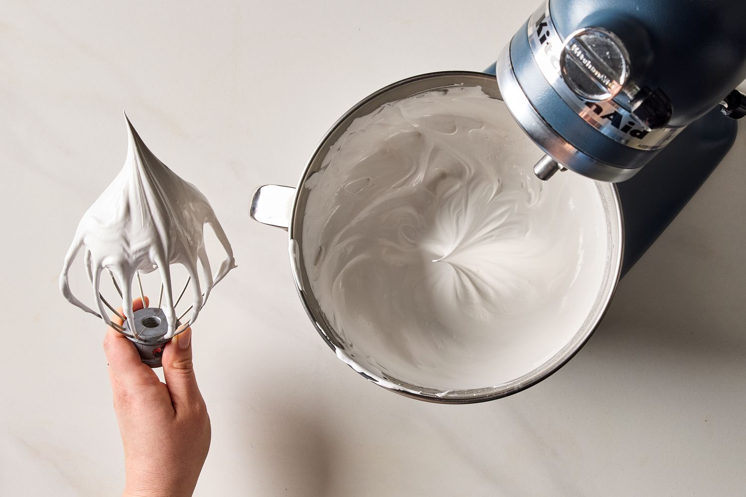 A mixing bowl on a stand mixer with marshmallow frosting and a hand holding the whisk attachment with frosting on it
