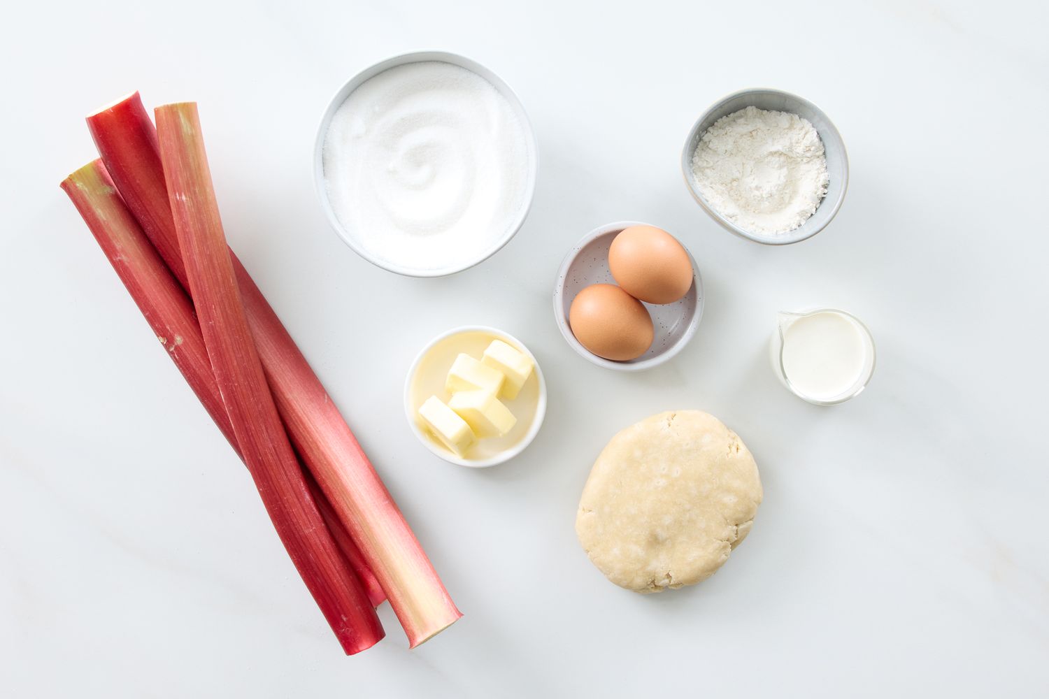 Ingredients for a rhubarb custard pie including rhubarb stalks sugar flour eggs butter milk and pie dough arranged on a surface