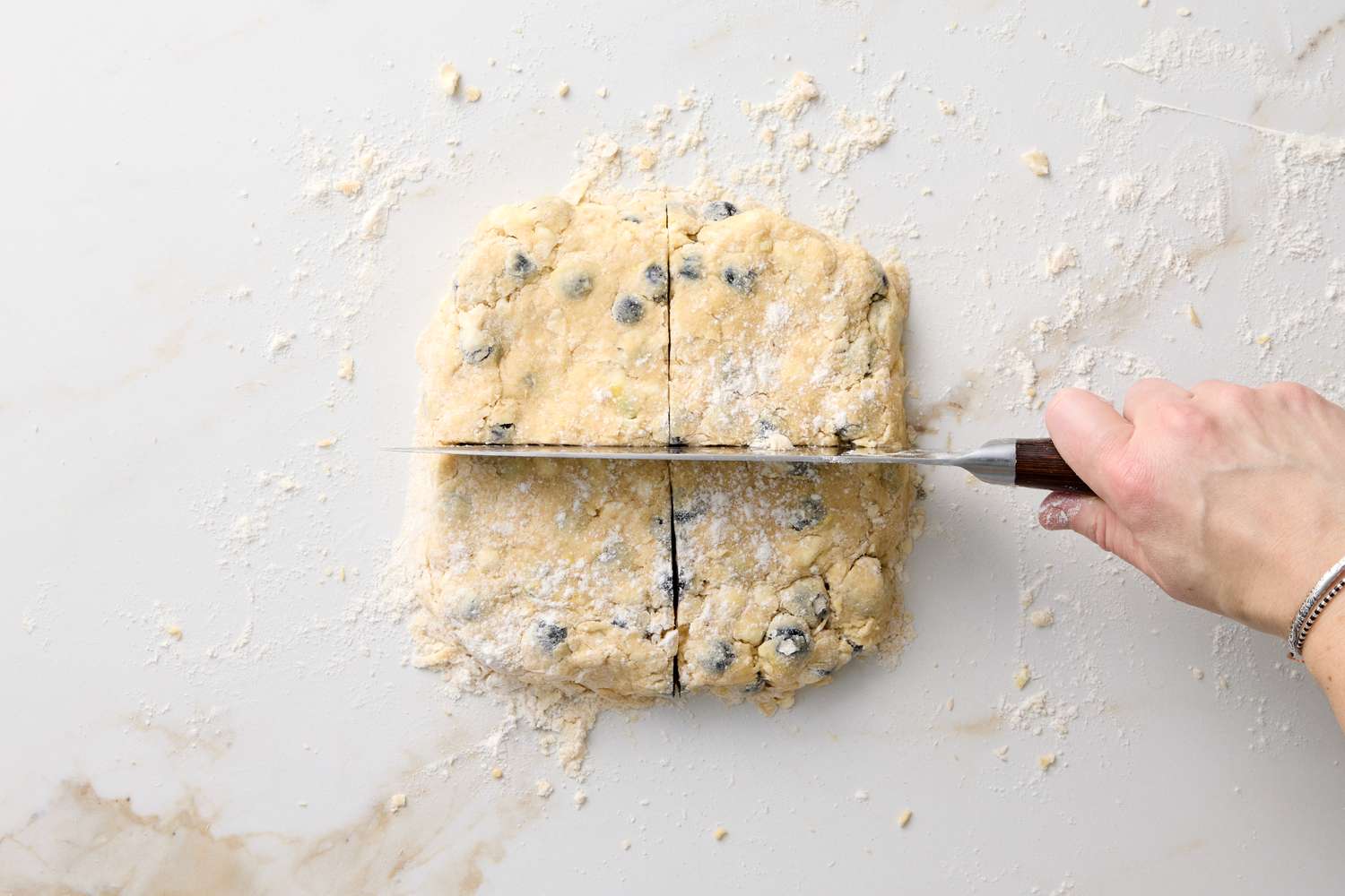 A hand holding a knife cutting blueberry scone dough into squares on a floured surface