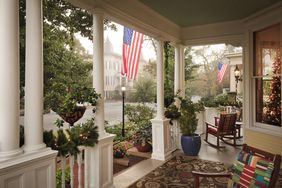 A porch decorated with seasonal greenery and American flags chairs and a patterned rug in the foreground treelined street in the background