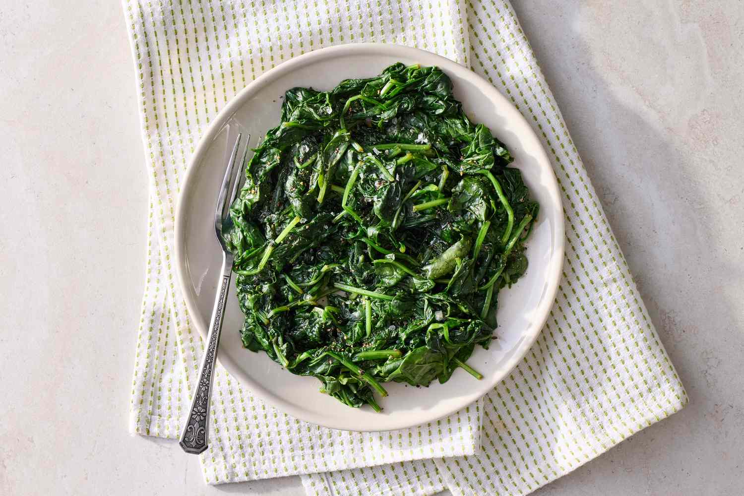 A plate of steamed spinach with a fork resting on the side placed on a patterned cloth