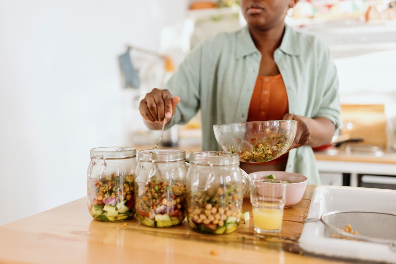 Person preparing salad jars in a kitchen scooping ingredients into glass containers on a counter
